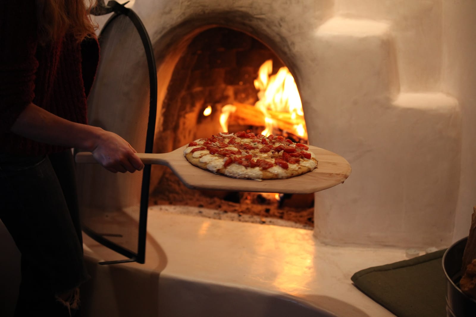 Nybagt pizza med tomat og mozzarella på en træspade foran en glødende brændefyret pizzaovn med åben ild
